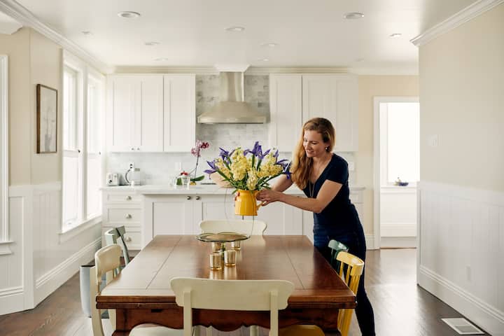 Female Host places colourful flowers on table with a white cabinet in the background.