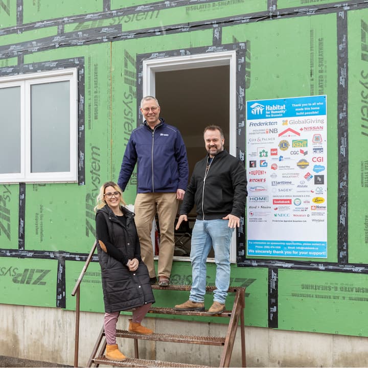 Three people smile standing on makeshift metal stairs in front of a house that is under construction.