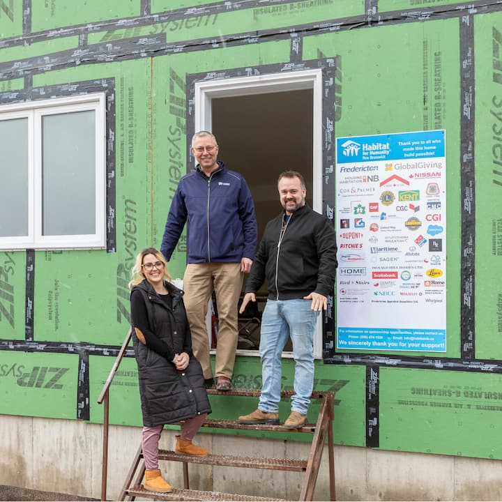 Três pessoas sorridentes em cima de escadas metálicas improvisadas em frente a uma casa em construção.