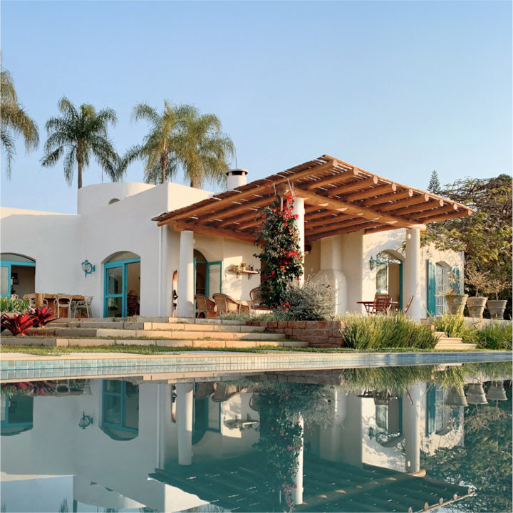 An image of an Airbnb home with an amazing pool. The image is of a gorgeous, white-washed home in some semi-tropical environment. A wooden overhang shades the patio area in front of the home’s modern pool.