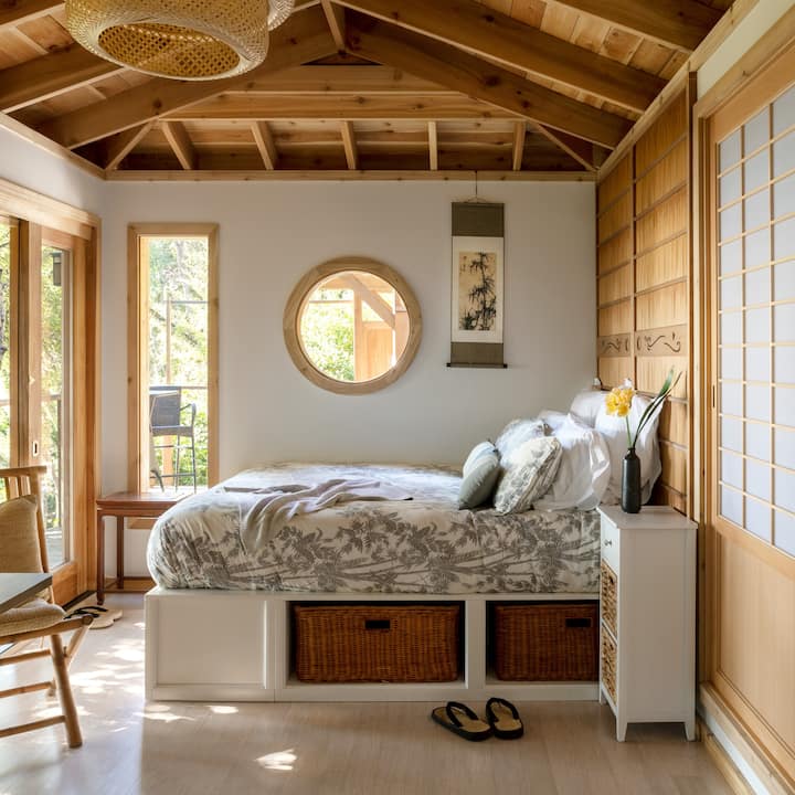 Une chambre à coucher d'inspiration japonaise avec des tons chauds et boisés, un lit joliment décoré et la lumière tamisée d'un arbre juste à l'extérieur de la fenêtre.