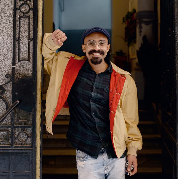 Um anfitrião feliz na porta de sua casa. O anfitrião usa óculos redondos, boné azul, jaqueta bege impermeável, camiseta xadrez verde e azul e jeans branco. Ele sorri com as chaves nas mãos.