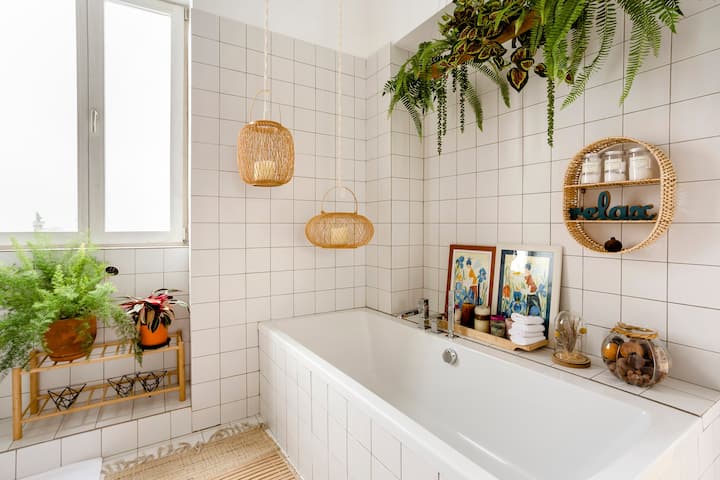 Lichte badkamer met grote vierkante witte tegels rondom een bad, decoraties van riet en muren met varens.