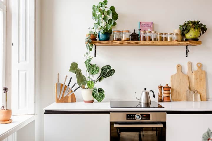 A bright and airy kitchen with neatly organized kitchenware: 3 small wood cutting boards lean against the wall, medium size glass jars filled with spices are featured on a wooden shelf, and 3 heathy plants sit atop the counter and shelf.