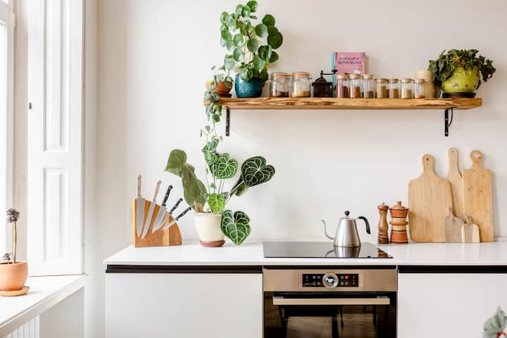 Una cucina luminosa e ariosa con utensili ben organizzati: tre piccoli taglieri in legno appoggiati al muro, barattoli di vetro di medie dimensioni pieni di spezie su un ripiano in legno e tre piante rigogliose appoggiate sopra il bancone e il ripiano.