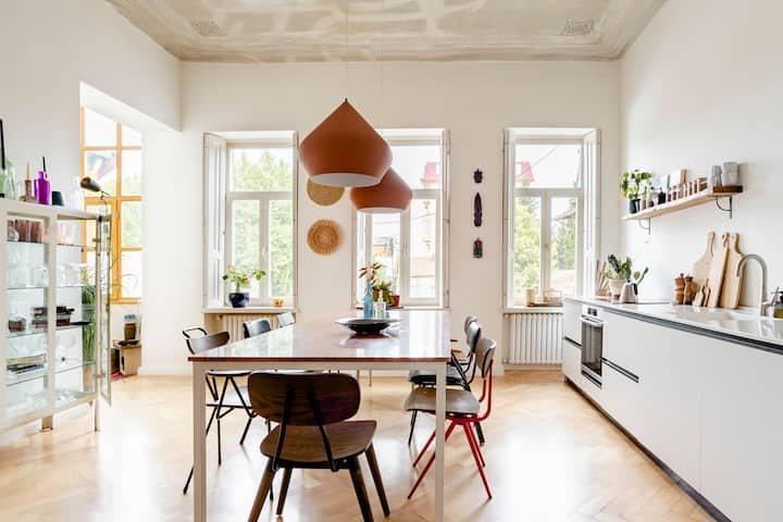 An wide image of a kitchen featuring the kitchen island on the right side, the dining table in the center, and a hutch filled with dish-ware on the left.