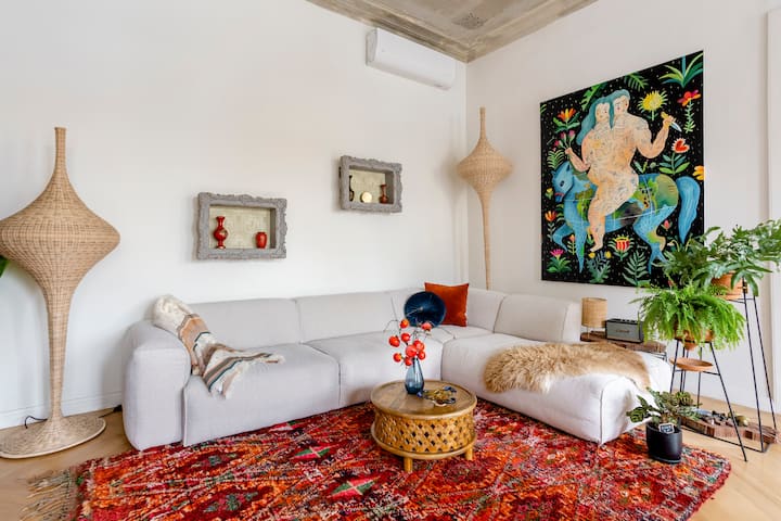 A beautifully style living room with a red Moroccan-styled rug, very large wicker lamps, a clean off white couch with a small sheep skin nicely styled on top, and plants to the right.