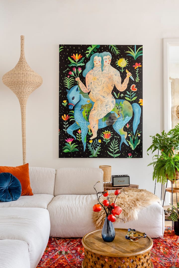 A vertical image of a close up of the living room details, including: a quirky celestial painting, with a very large wicker lamp, an off-white couch with colorful pillows, and plants to the right.