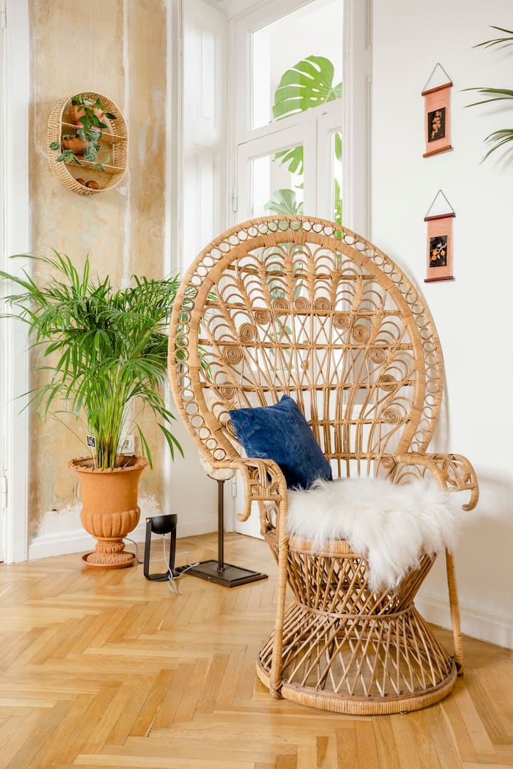 Een verticale afbeelding van een hoek van de kamer. Daar staan een rieten stoel met een witte schapenvacht en een blauw kussen, en een grote plant in een terracotta pot.