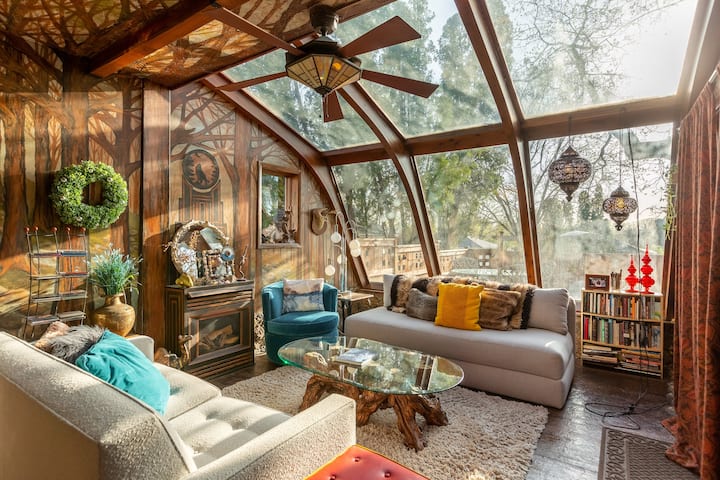 A living room with a whole wall made of floor to ceiling windows and wooden panels that look like trees. The afternoon sunlight shines in.