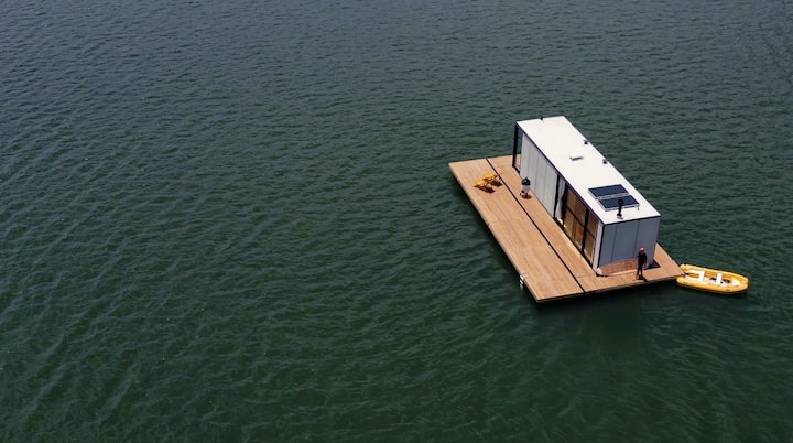An image of a  modern floating house on a lake, with a wooden deck, large glass windows, solar panels on the roof, and a yellow kayak beside it.