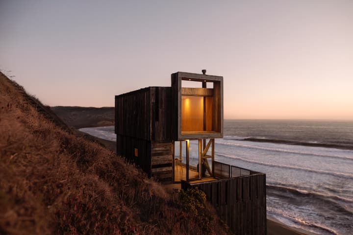 An image of a modern wooden cabin lit from within stands on a coastal cliff overlooking the ocean at sunset.