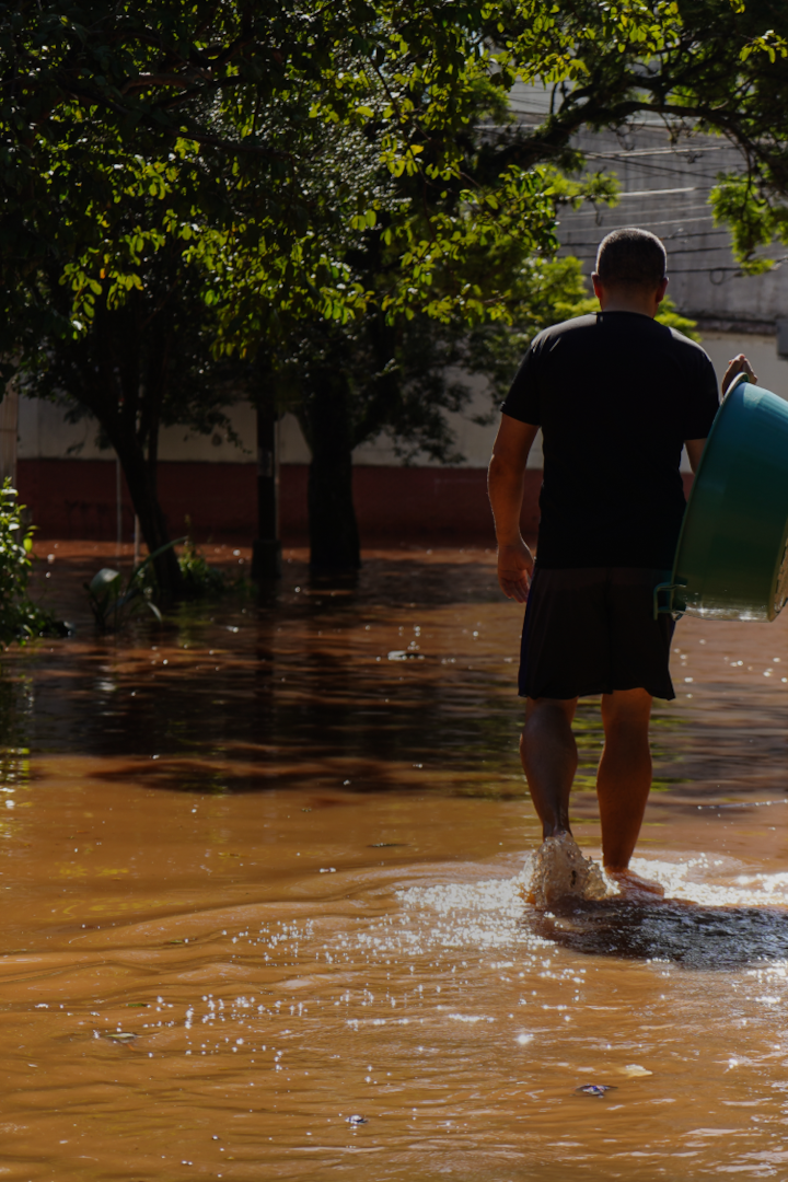 Uma pessoa com cabelos escuros bem curtos se afasta, carregando uma banheira de plástico por uma rua inundada.