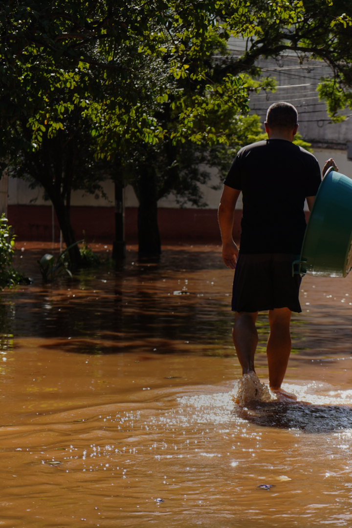 A person with shorn dark hair walks away, carrying a plastic tub through calf-deep muddy water on a flooded road.