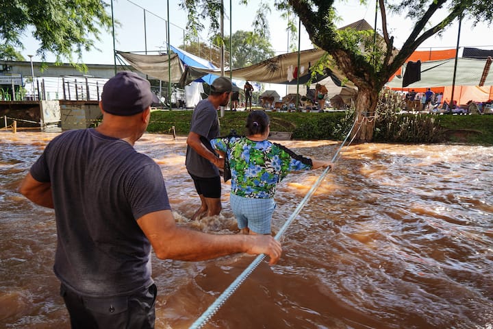 Three people cross fast-moving, knee-deep, muddy water, holding a rope and each other.