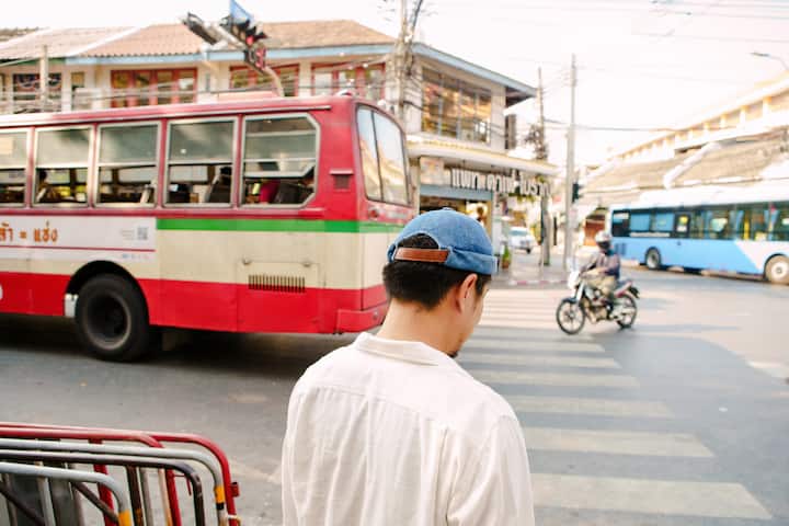 A young man wearing a hat crosses a road in a city. The camera is behind him