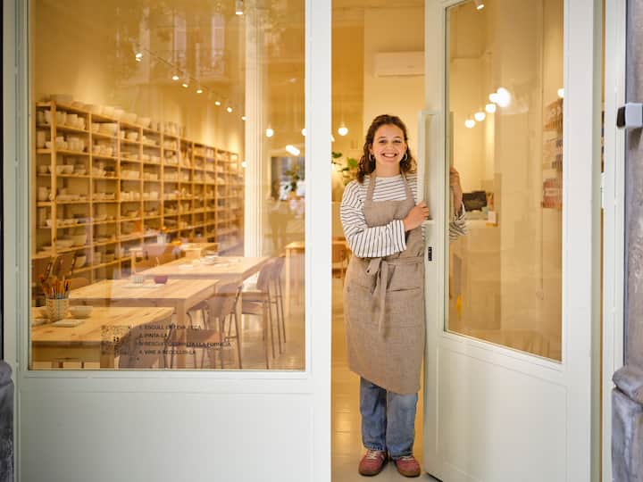Una joven abre la puerta de un estudio de cerámica. Lleva un delantal y sonríe con amabilidad.