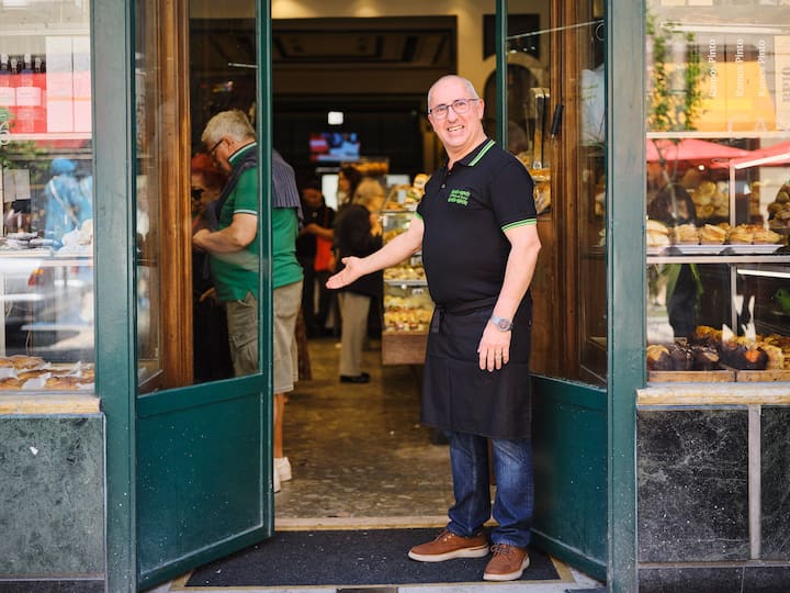 Un hombre da la bienvenida a viajeros en su pastelería.