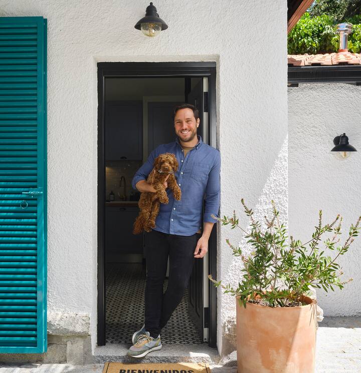 Jonathan, anfitrión en Airbnb, recibe a los huéspedes con la puerta abierta y su perro en brazos. Fuera brilla el sol, y su casa parece luminosa y acogedora.