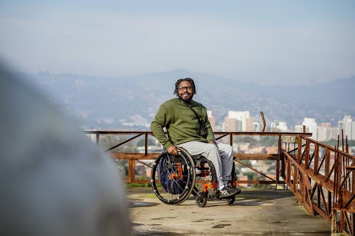 車椅子に座っているヴァンデルソン・チャベスが、展望台からブラジルの街を眺めながら微笑んでいます。背景には山の景色が広がっています。
