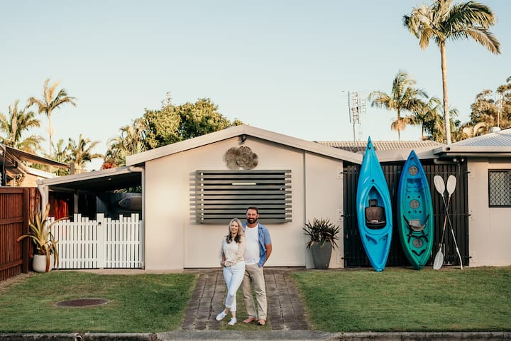A couple stands in front of a small beachside house with kayaks and paddles leaned against the wall, surrounded by palm trees.