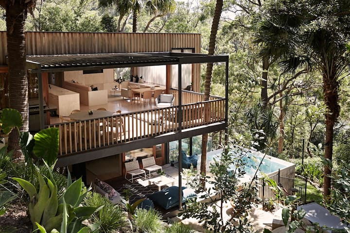 The image shows a multi-level modern home nestled within a lush, green forest. The upper level features an open-plan living space with wooden furniture and large sliding doors that open onto a spacious balcony, offering an indoor-outdoor flow. Below, a cozy outdoor lounge area is visible, leading to a sleek, rectangular pool surrounded by tropical plants. The home blends seamlessly with its natural surroundings, creating a tranquil, resort-like atmosphere.