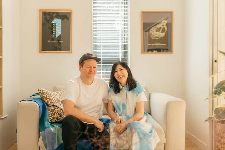 A smiling couple sitting on a couch in a bright, modern living room with framed artwork on the wall and soft natural light coming through a window.