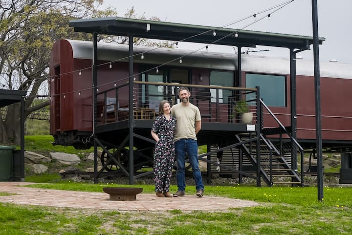 Couple standing lovingly outside maroon train carriage airbnb