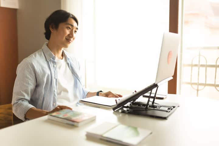 Person sitting at a table working on a laptop, with books and papers spread out in front of them in a bright room.