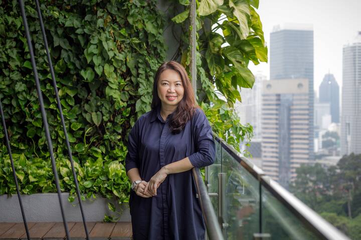 Individual in a dark blue dress on a balcony with lush greenery and a view of tall buildings in the background.