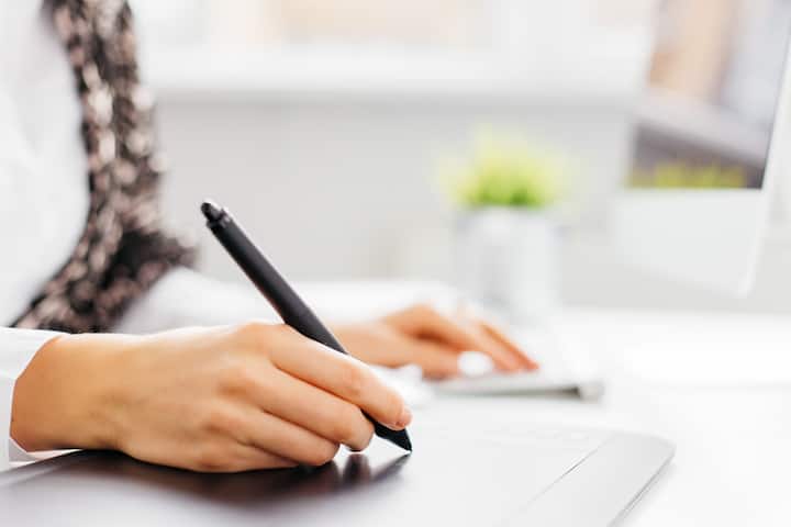 Close-up of a person using a digital pen on a tablet, with a blurred computer and plant in the background.