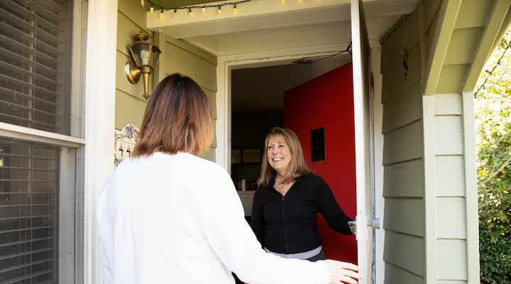 A host welcoming a guest at the front door of a house, with warm lighting and a bright red door.