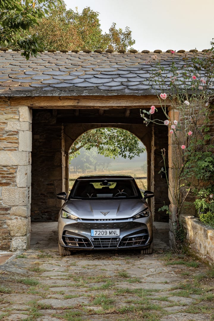 Un coche Cupra plateado está estacionado en la entrada delantera de un garaje rústico de piedra con techo de pizarra. El coche está parcialmente oculto por el arco, y a la derecha de la imagen se pueden ver flores rosas y vegetación que le agregan un toque natural a la escena. El fondo más allá del garaje muestra un paisaje tranquilo, iluminado por el sol y con árboles.