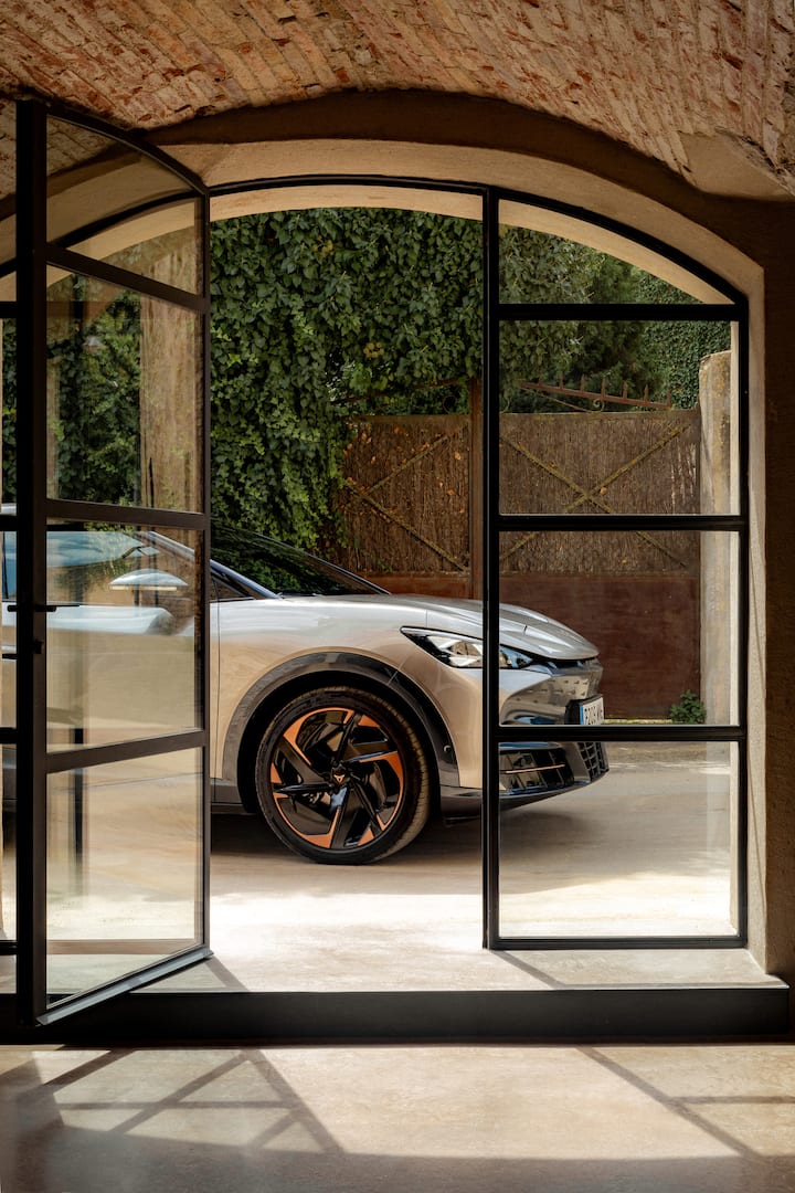 Coche eléctrico de Cupra aparcado visto desde el interior de una puerta de cristal arqueada con enredaderas en el fondo.