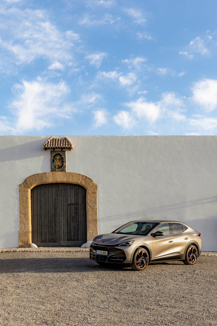 Un coche eléctrico de Cupra está estacionado afuera de un edificio tradicional andaluz de color blanco, con una gran puerta de madera y una maceta con una planta.