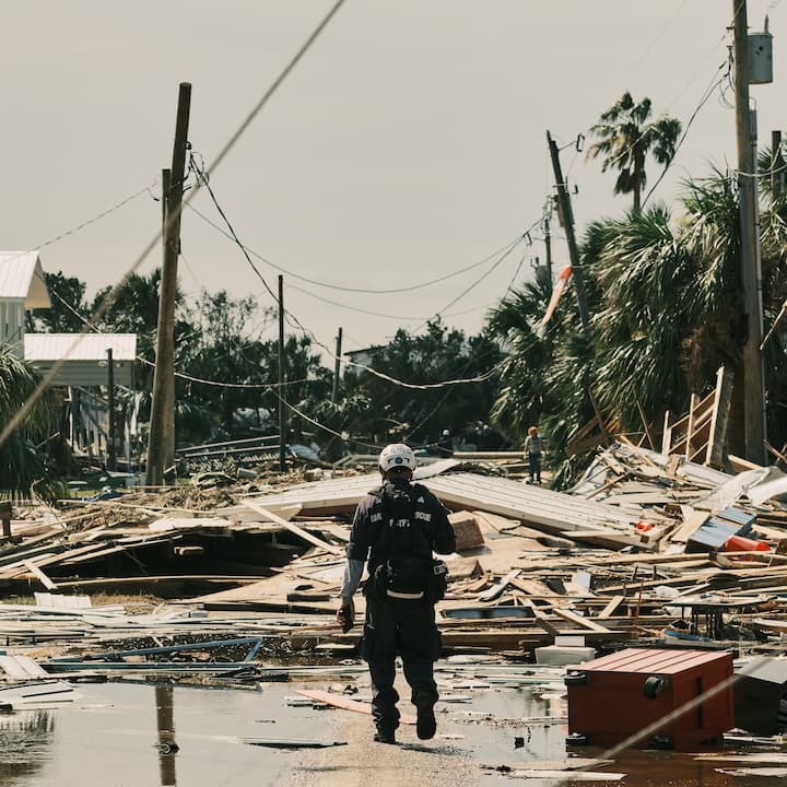 Rescue worker walking through hurricane debris with collapsed houses and fallen trees in the background.