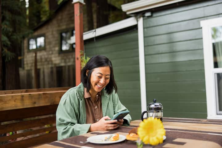 Woman sitting at a picnic table outside smiling and looking at her phone.