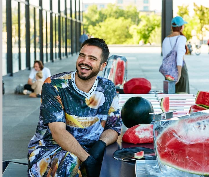 A man sits at a table on a terrace, smiling. He’s surrounded by a handful of watermelons.