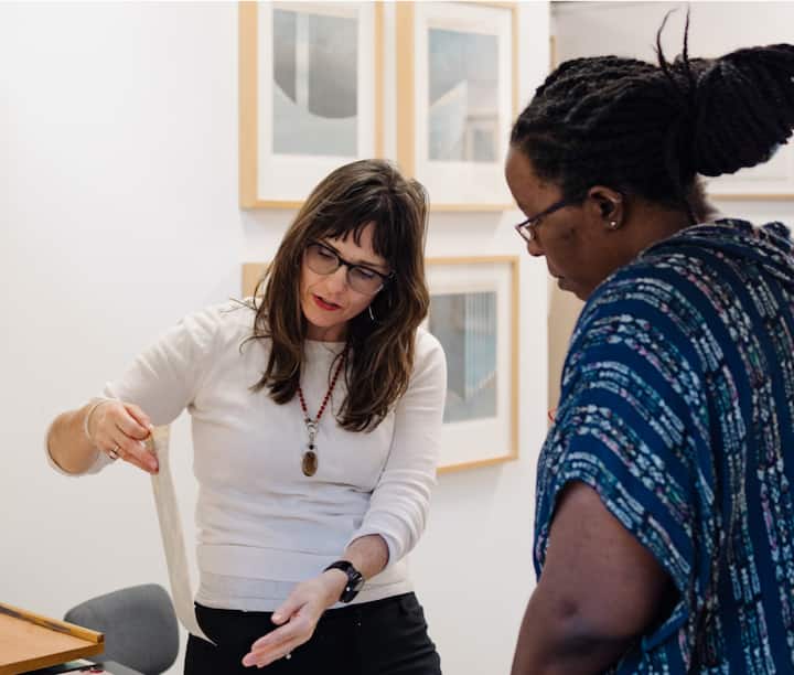 A woman stands at a desk in a white art studio, holding up a print to show to a patron.