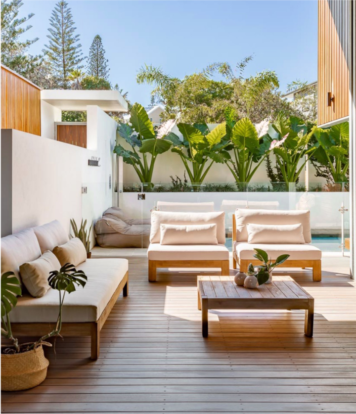 Uma casa moderna com um quintal privativo cheio de plantas tropicais altas. Uma espaço de convivência sombreado leva a uma piscina com uma cerca de vidro.