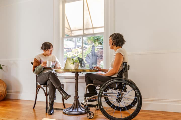 Two women speak by a sunny window. One woman is a wheelchair user.