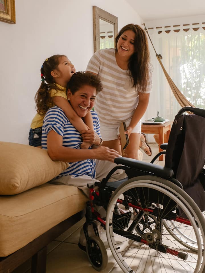 Trois générations d'une famille sourient et rient dans un logement Airbnb accessible. Un fauteuil roulant se trouve devant le membre le plus âgé de la famille.