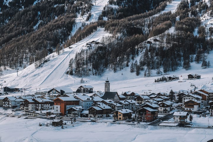 Verschneites Dorf am Fuße eines bewaldeten Berges mit Skipisten, mit Gebäuden mit dunklen Dächern und einem zentralen Kirchturm.