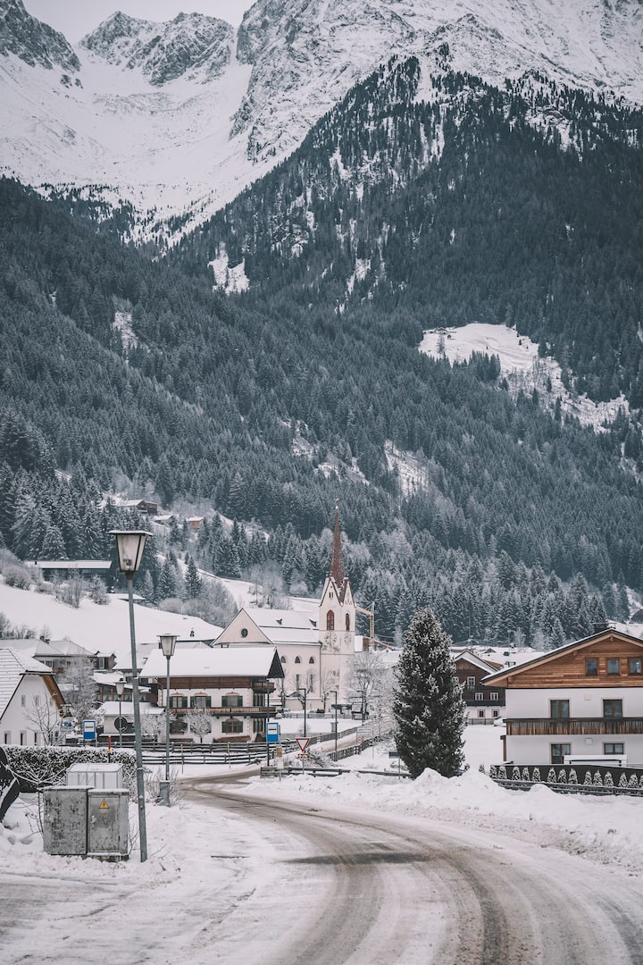 Kleines verschneites Dorf mit einer Kirche mit hohem Kirchturm, umgeben von schneebedeckten Bäumen und Bergen.