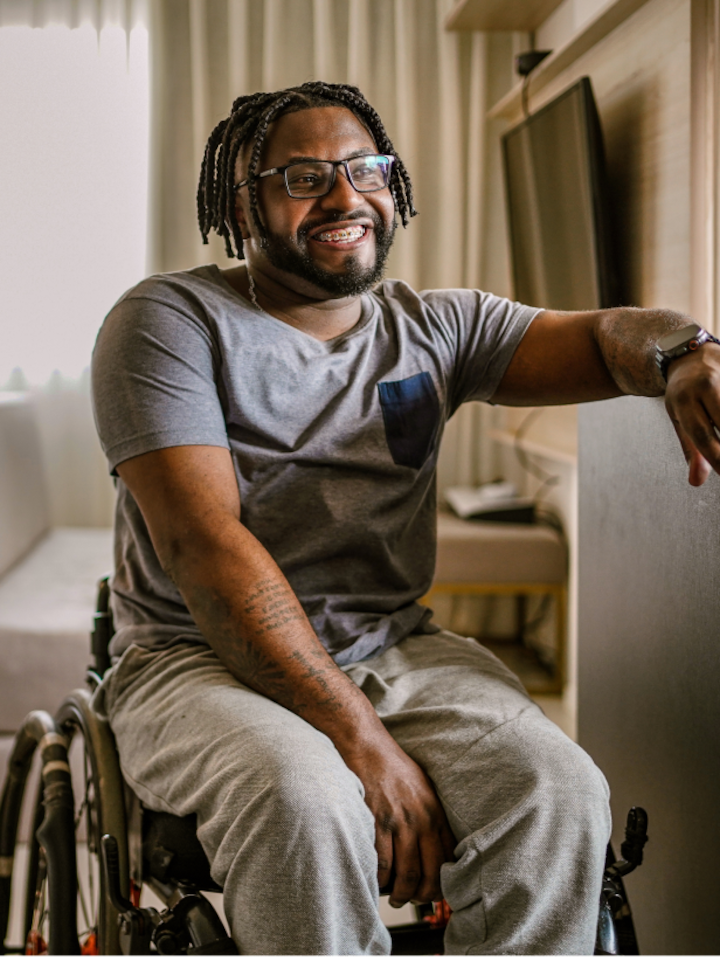 A man sits in a wheelchair smiling with his arm resting on the counter, in a room with a TV and a desk in the background.
