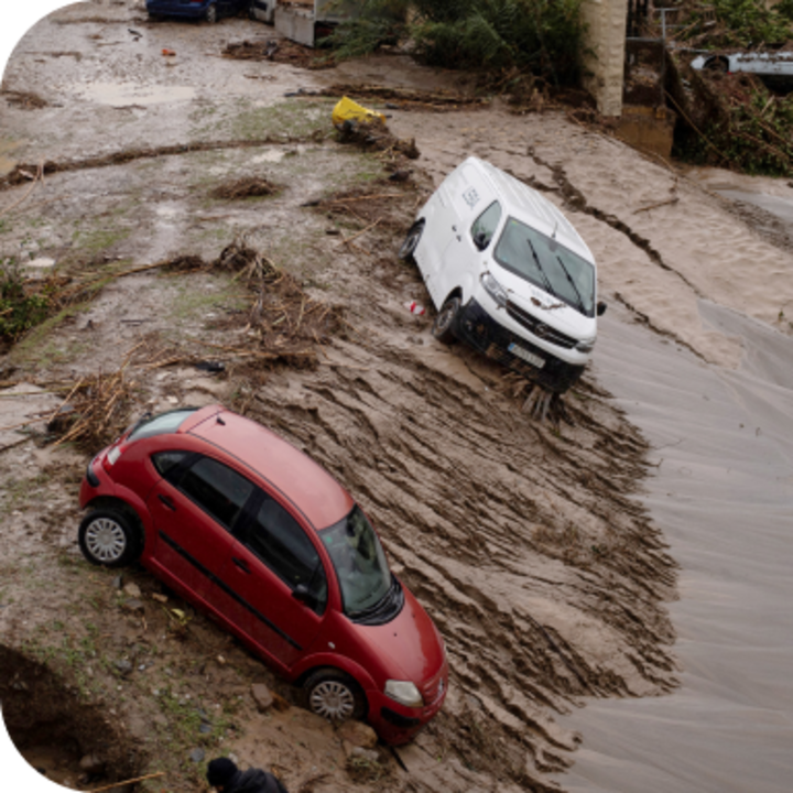 A flood-damaged red car and white van lean on muddy ground above flood waters.
