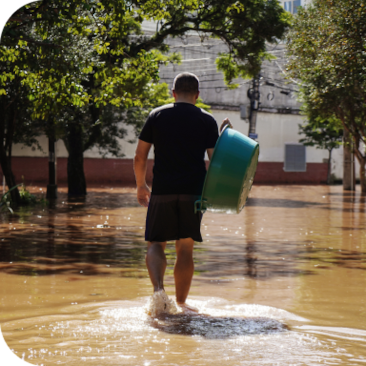 A man carrying a green bucket wades through muddy water in front of trees and a telephone pole on a sunny day.