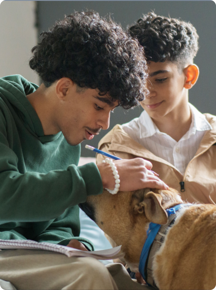 Two boys sit next to each other on a bed, one of them petting a dog while holding a notebook and a pen.