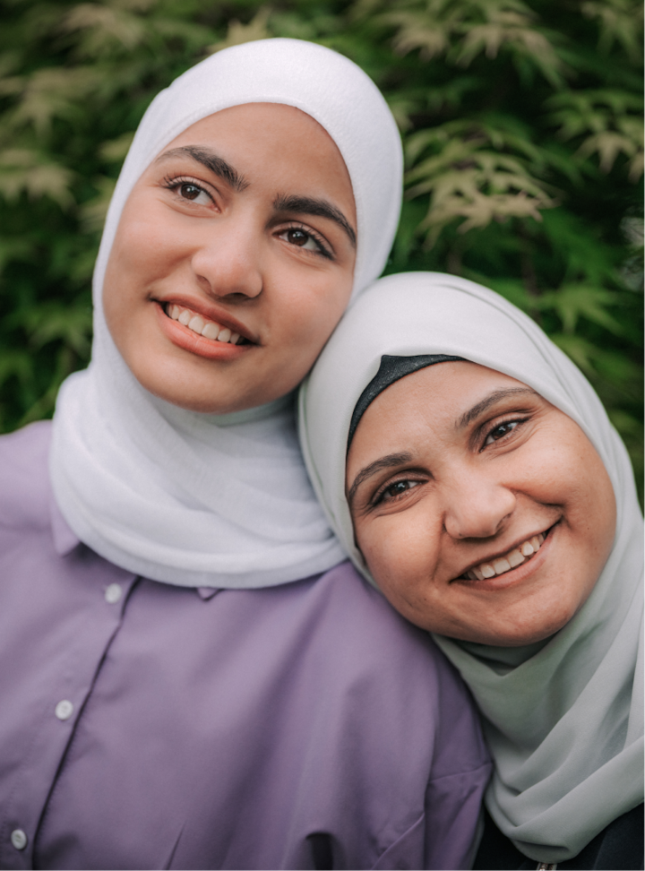 Two women in light-colored hijabs lean into each other smiling, with leafy greenery in the background.