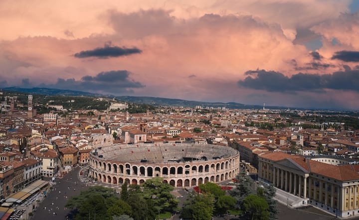 Luftaufnahme von Verona, Italien, mit dem antiken römischen Amphitheater Arena di Verona, im Hintergrund ein rosa-orangener Sonnenuntergang.